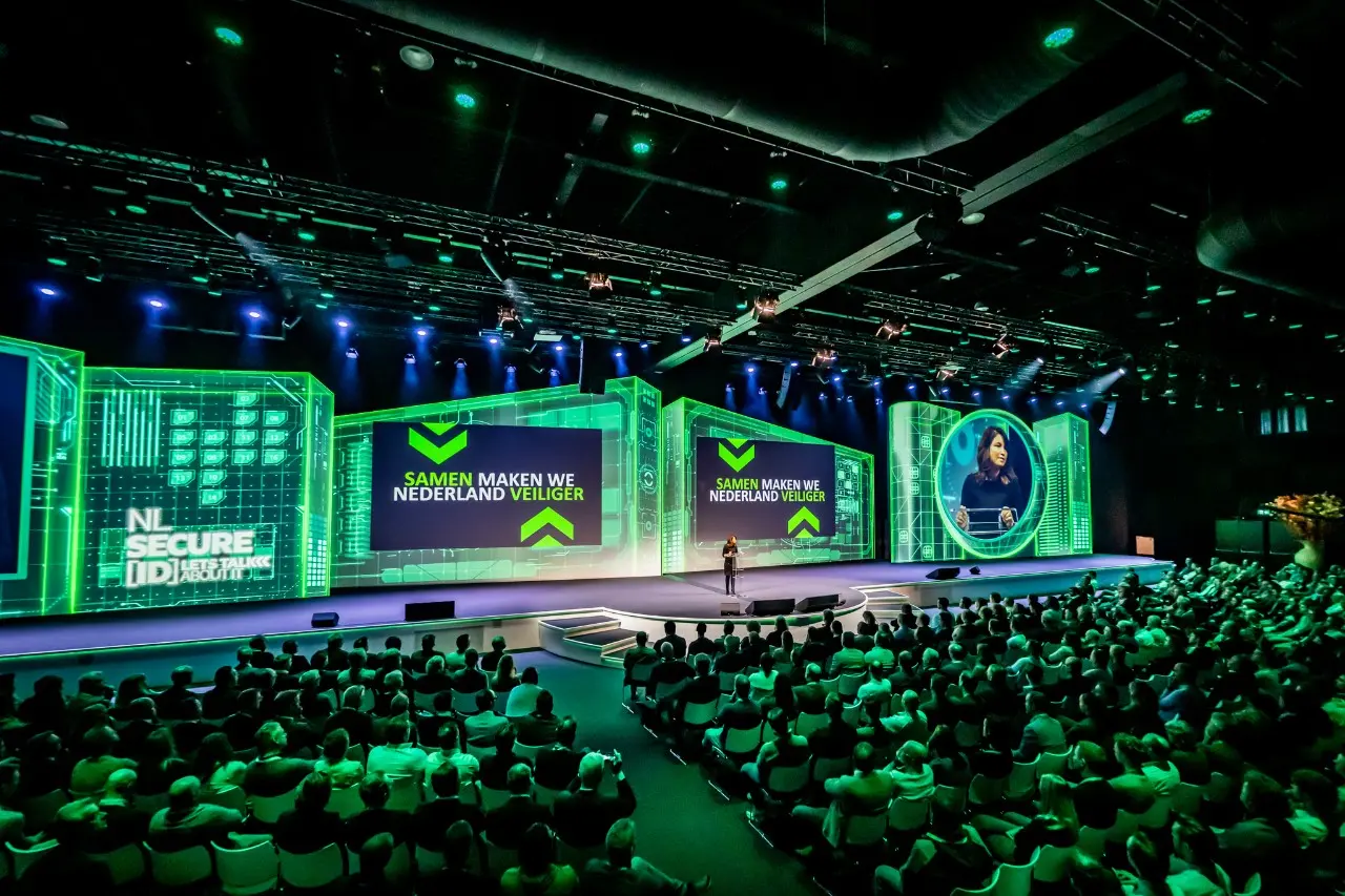 Zaal met mensen kijkt naar een podium toe. Op het scherm is 'Let's talk about progress' te zien. 