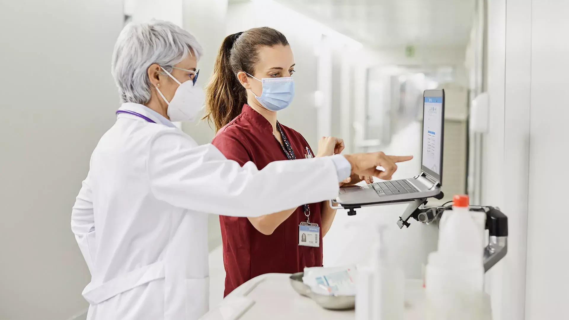 Twee vrouwelijke artsen met mondkapjes kijken naar een laptop in een ziekenhuisgang
