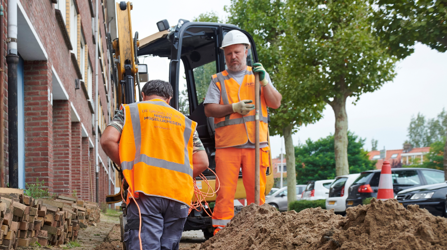 Twee mannen in oranje hesjes graven een gat in de stoep. Ze leggen een glasvezelverbinding aan.