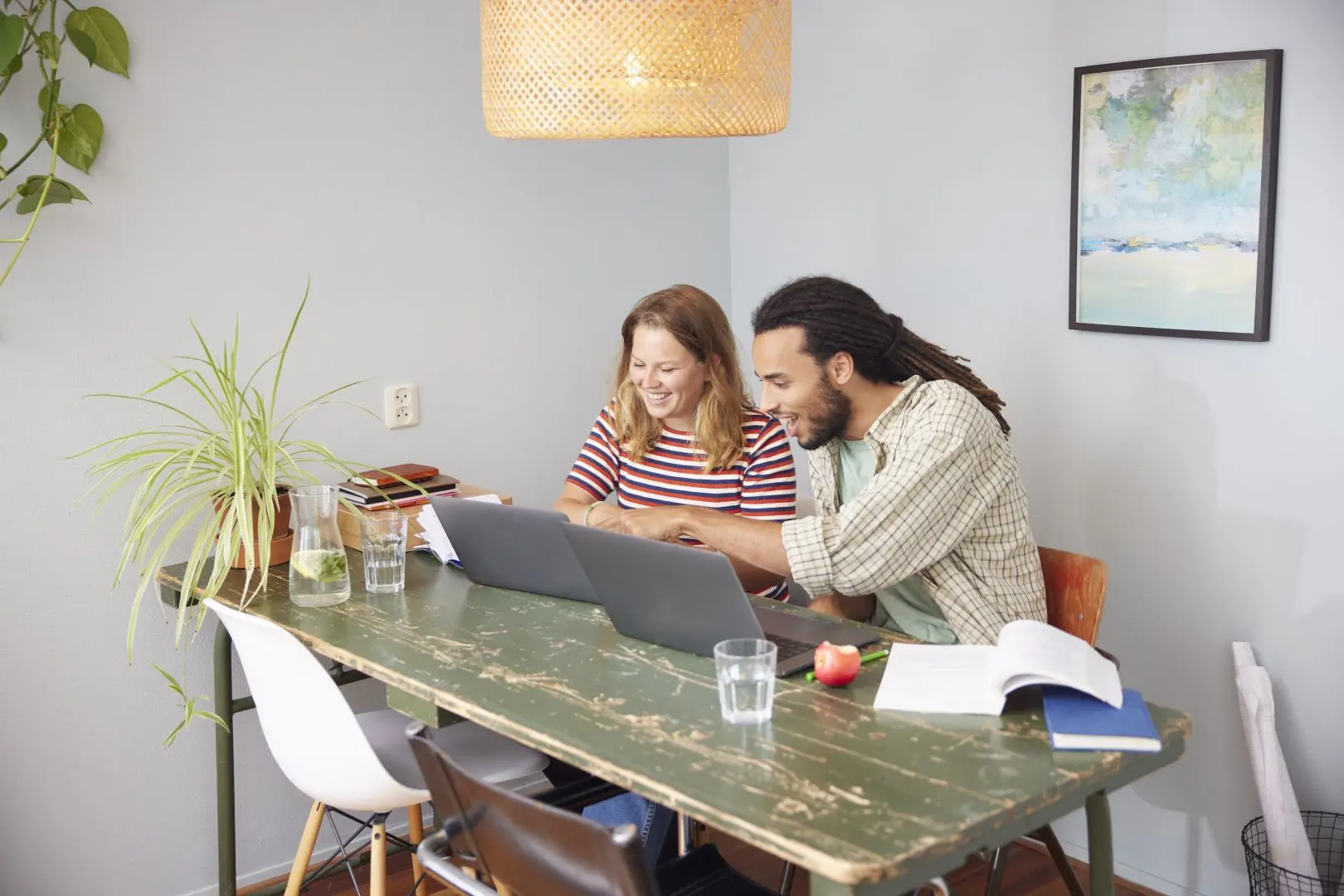 Lachende jonge man en vrouw aan een eettafel, elk achter een laptop