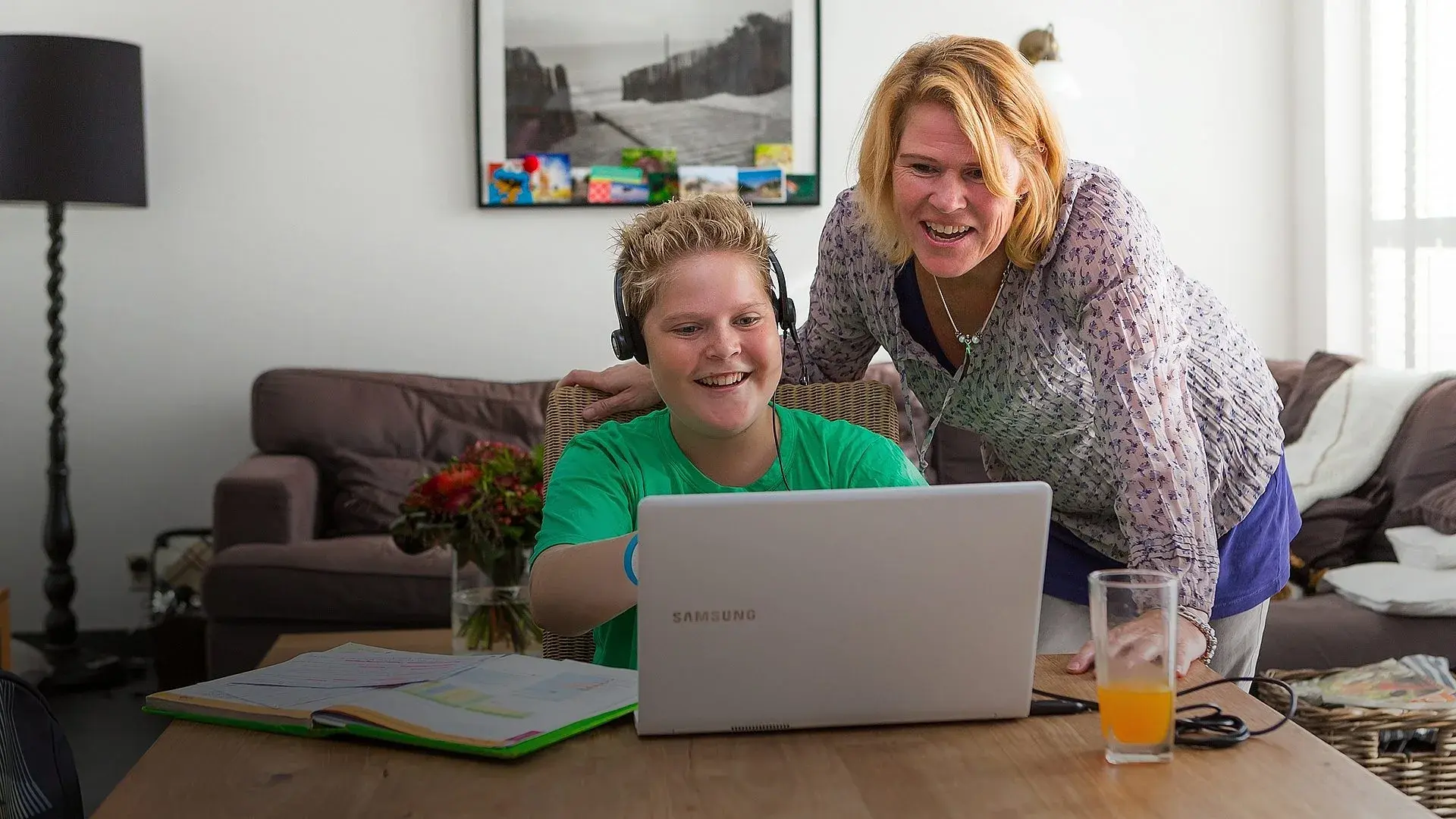 Moeder kijkt bij zoon over de schouder mee die vanuit huis actief deelneemt in de klas via De Klasgenoot ICT-apparatuur