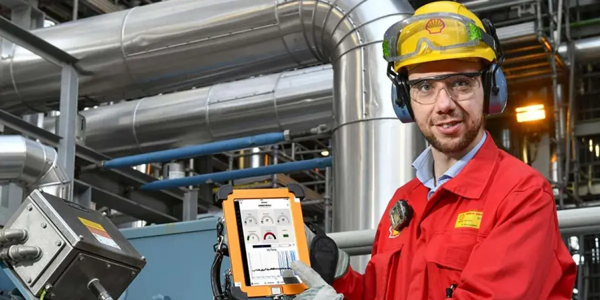 Een man in overall van Shell met helm en oorbeschermers op laat in een fabriek zijn mini-tablet zien