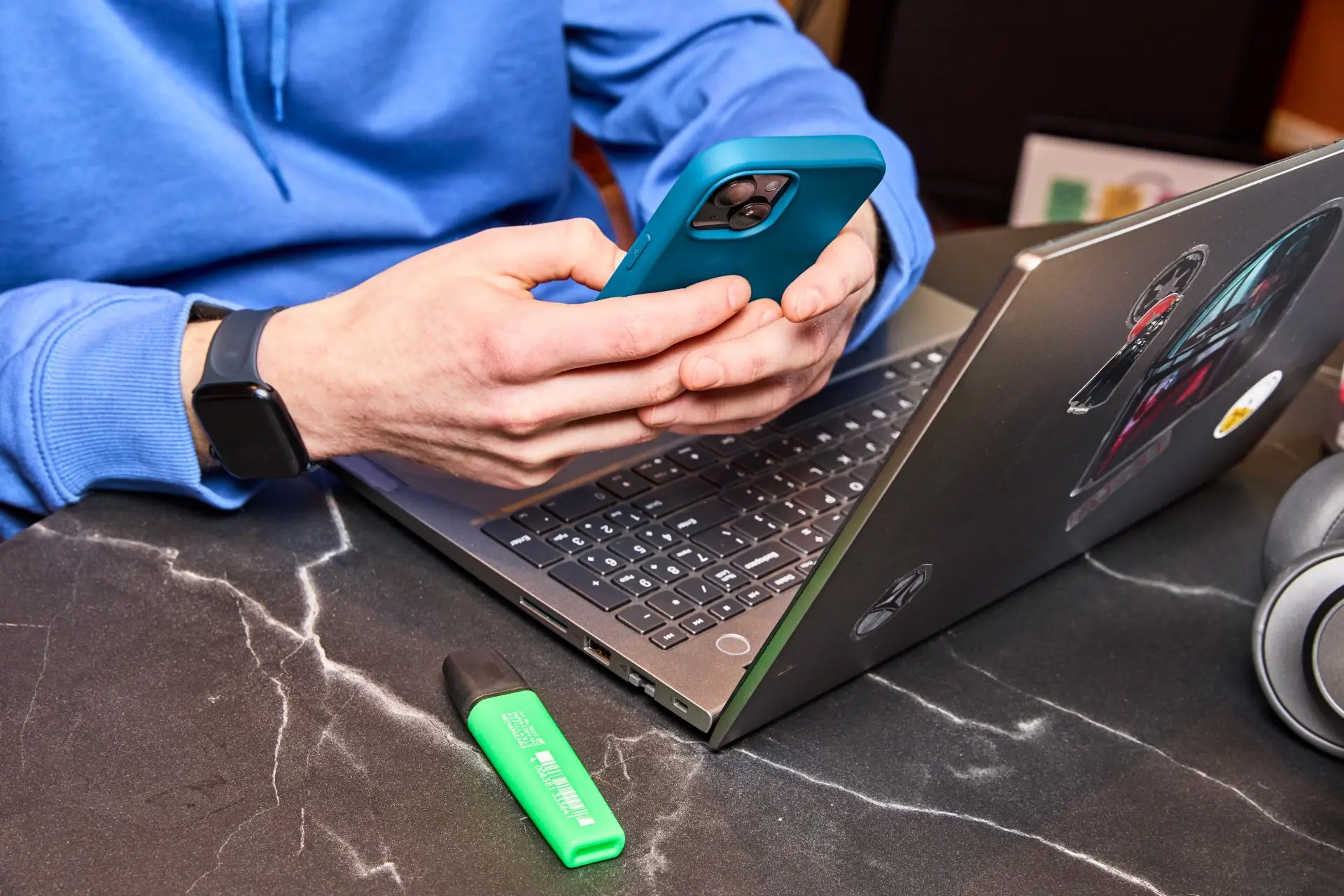 Persoon met mobiel in handen achter een laptop en marker op bureau