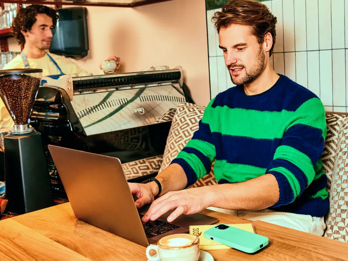 Een man aan het werk op zijn laptop in een koffiezaak. Op de achtergrond werkt een barista.