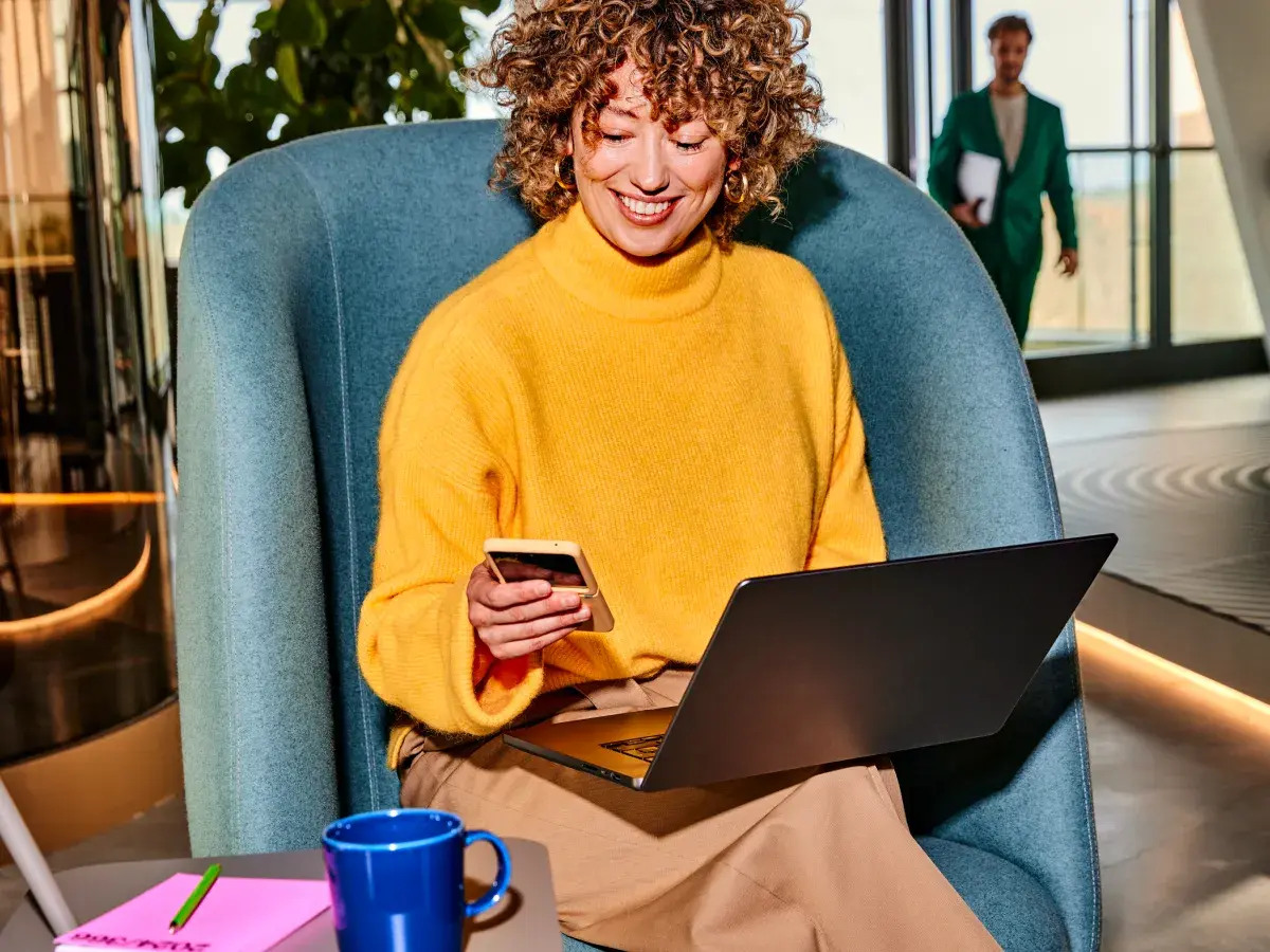 Vrouw zit op blauwe stoel met laptop op schoot en mobiel in hand 