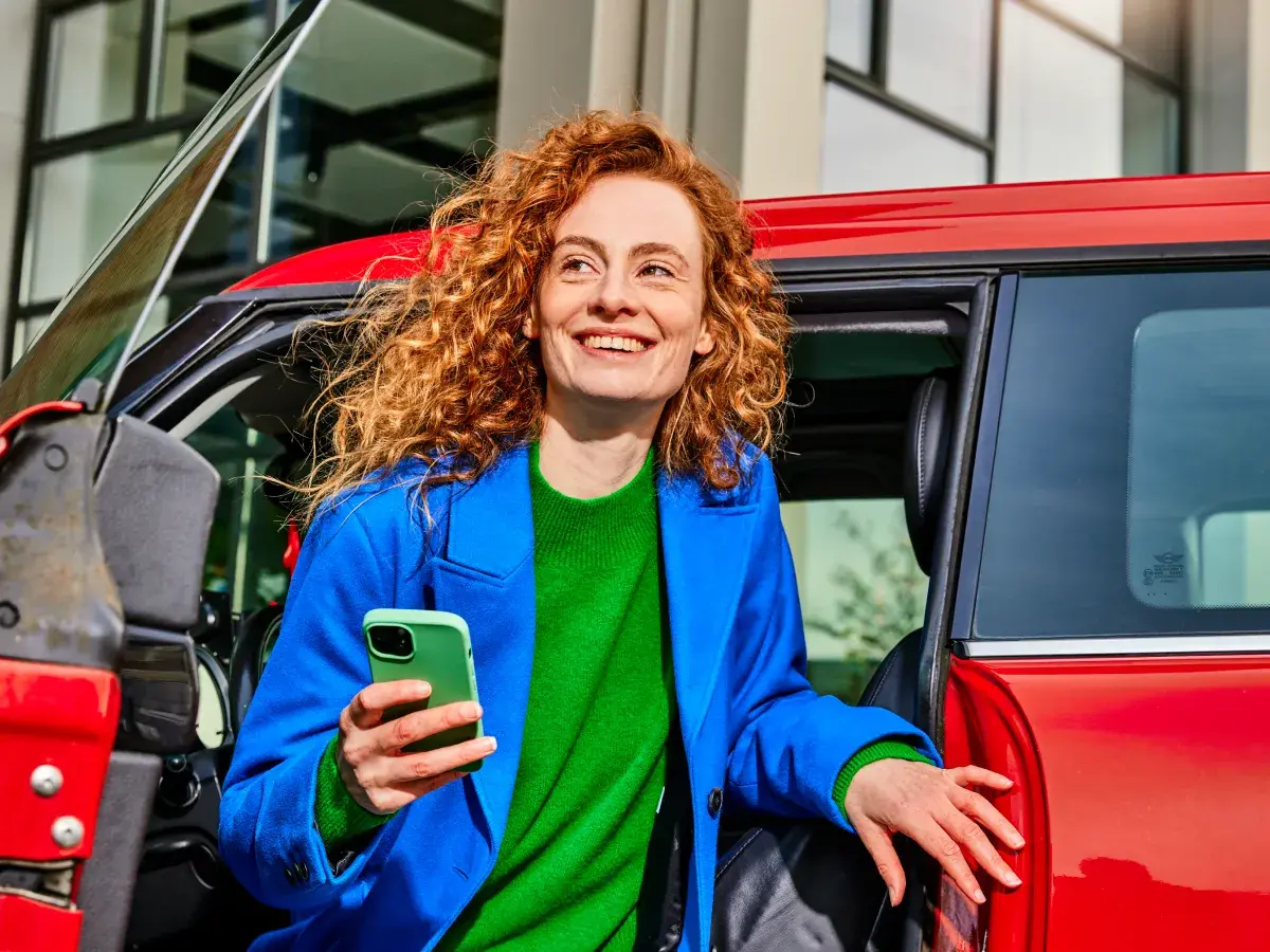 Vrouw stapt uit rode auto, lacht en heeft haar telefoon in de hand