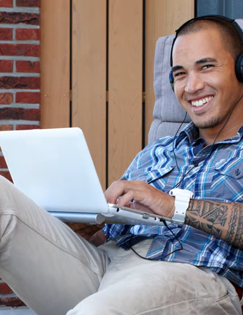 Een man zit buiten met een koptelefoon op. Op zijn schoot staat een laptop. Hij lacht in de camera.