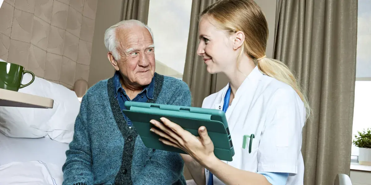 Een arts en een oudere man zitten samen op bed. De arts heeft een iPad in haar handen.