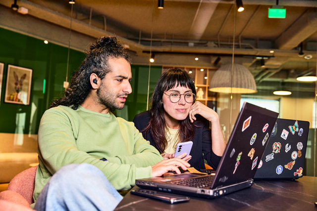 Man en vrouw op een kantoor achter hun laptop. De vrouw houdt een telefoon vast