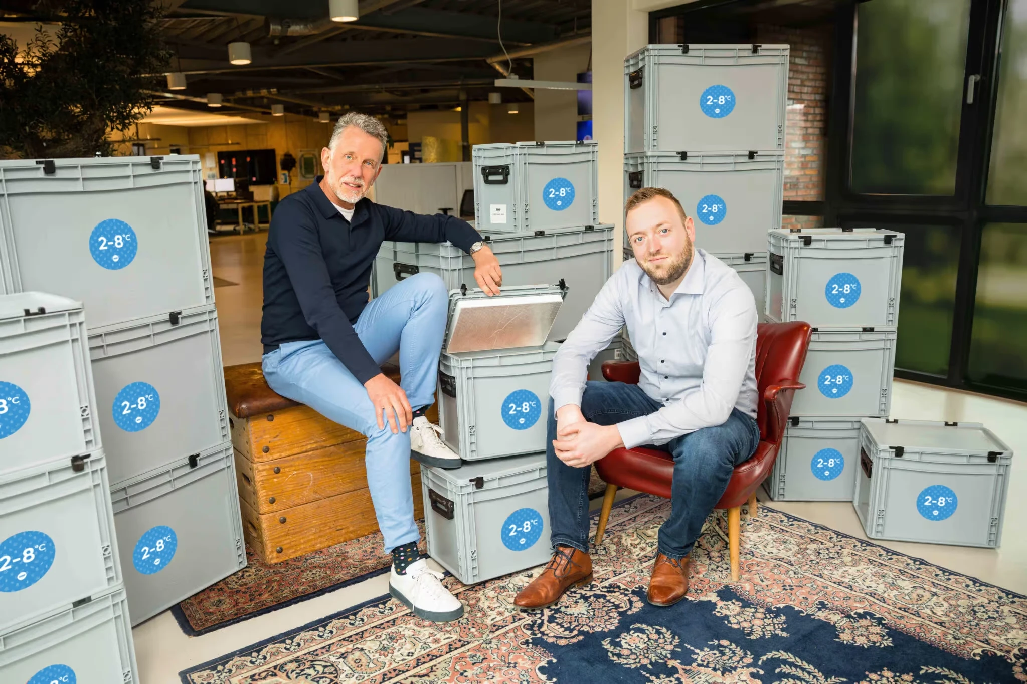 Twee medewerkers van AMP zitten bij medicijntransportboxen en kijken vriendelijk naar de camera 