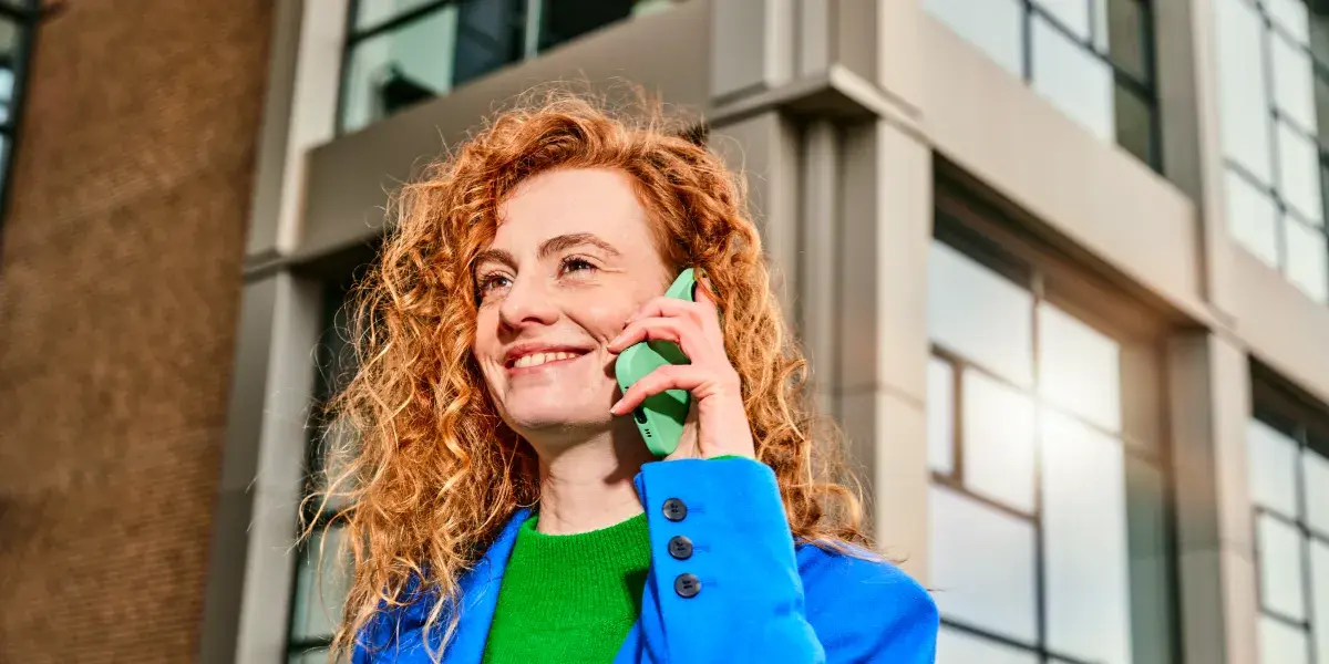Een vrouw met rood krullend haar staat buiten en belt met haar mobiele telefoon.