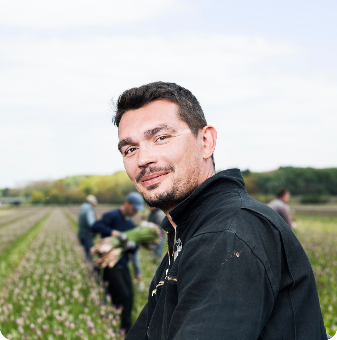 Man kijkt in de camera. Op de achtergrond werken mensen op het land.