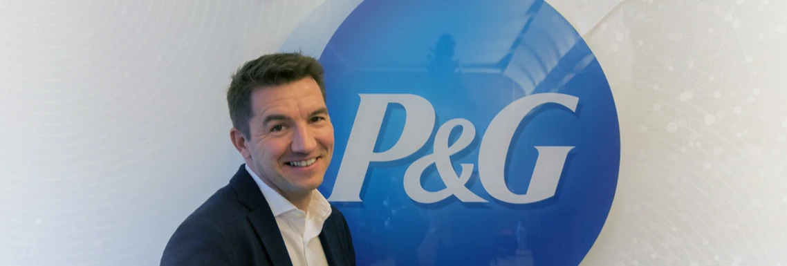 Gian De Belder, Technical Director, R&D Packaging Sustainability smiling towards the camera, wearing white shirt and a dark suit. He is standing in front of a P&G logo.