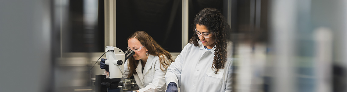 Two women in lab coats and safety glasses work in a laboratory, one looking into a microscope and the other manipulating molecular models.
