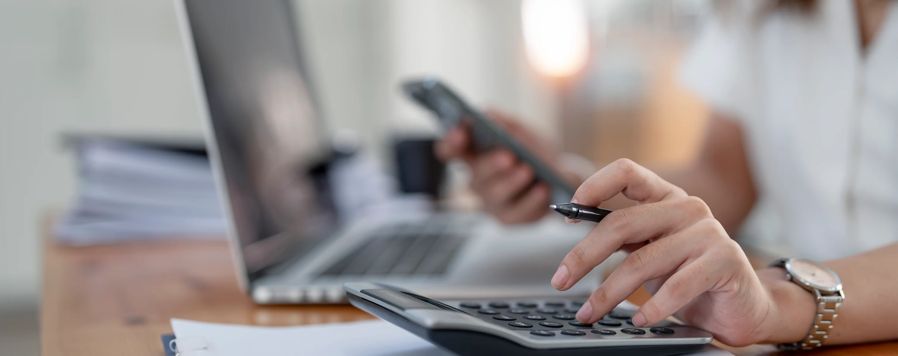 Eine Person nutzt einen Taschenrechner, während sie gleichzeitig ein Smartphone in der Hand hält; im Hintergrund steht ein Laptop auf einem Schreibtisch.