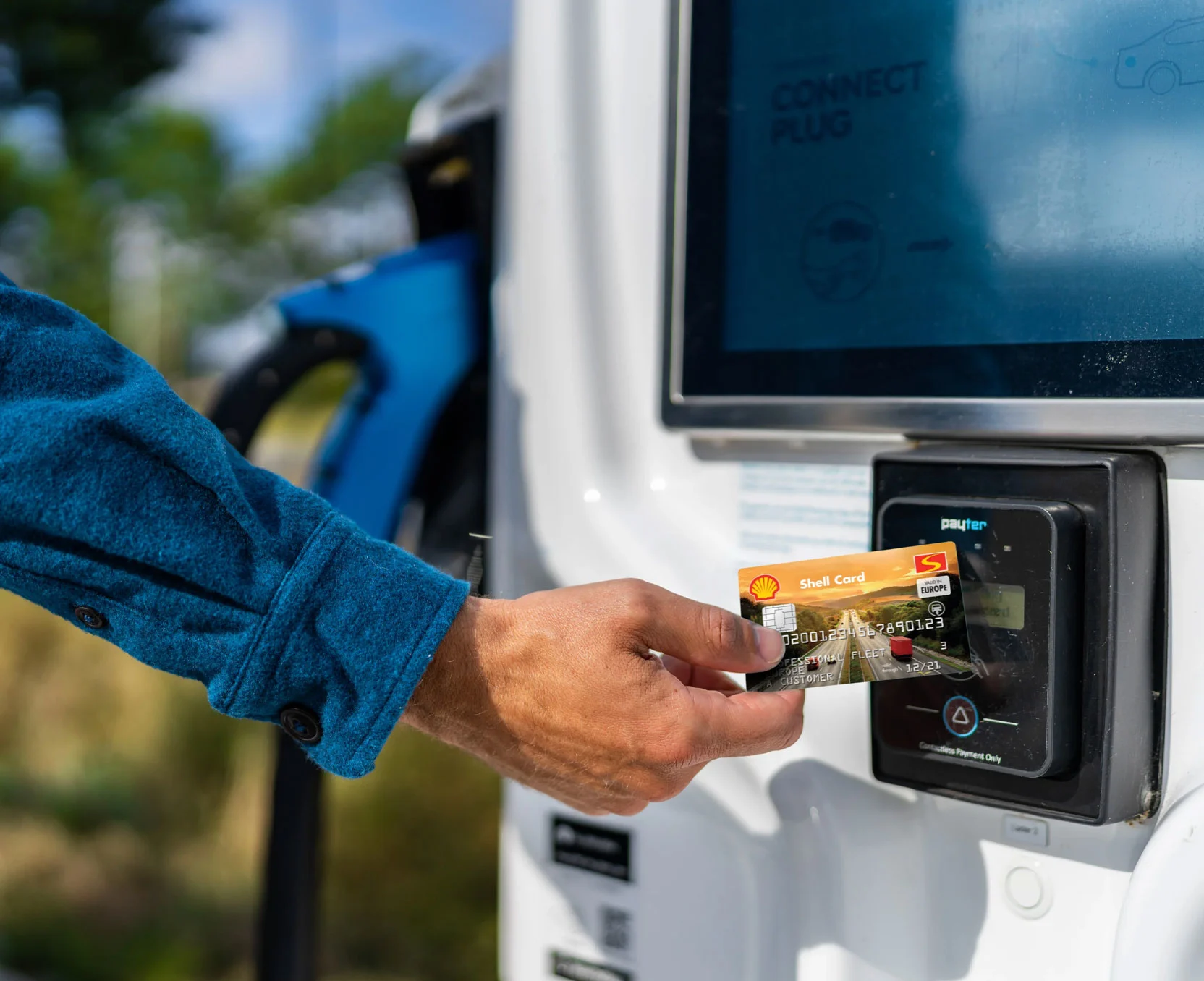  Ręka trzyma kartę Shell przed terminalem płatniczym przy stacji ładowania.