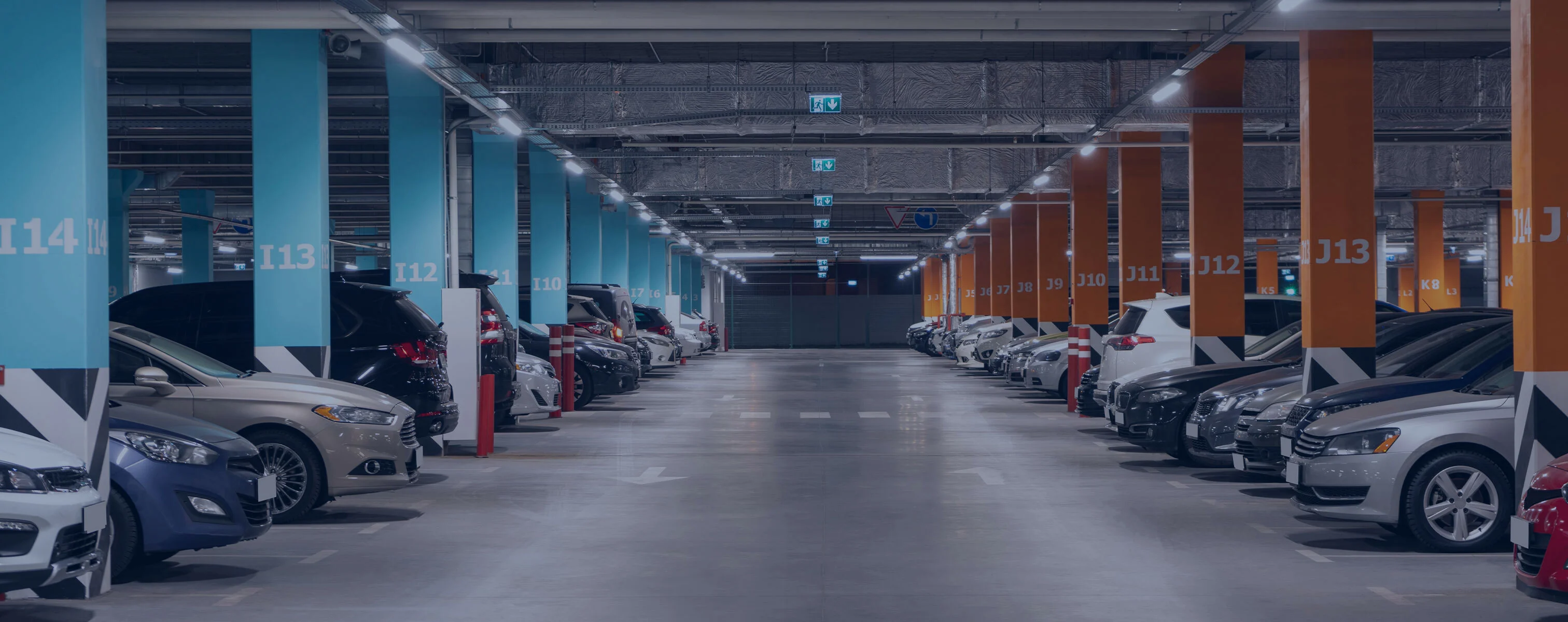 Parking moderne bien éclairé avec des places marquées, dans lequel de nombreuses voitures sont garées des deux côtés entre des rangées de colonnes colorées.