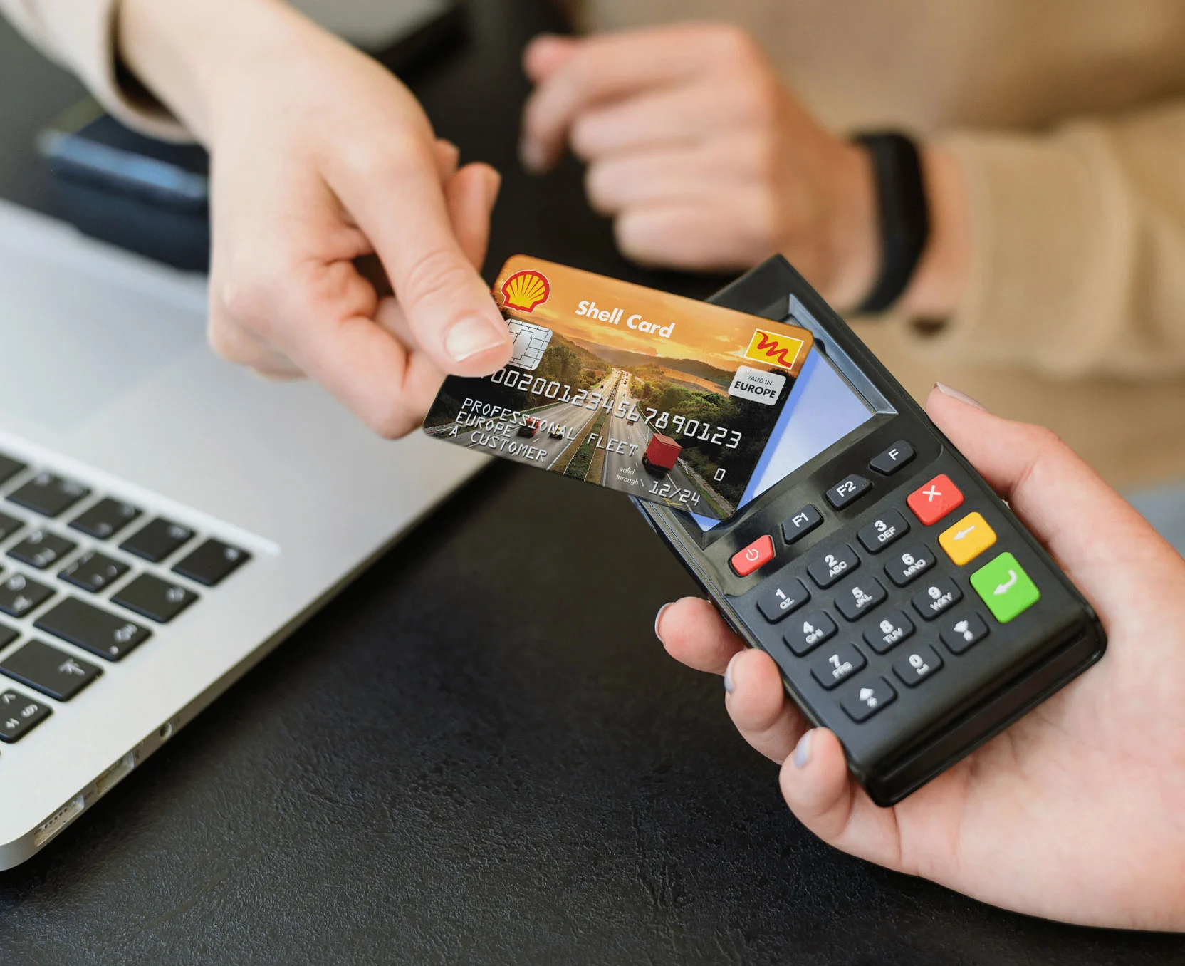 Osoba płaci kartą Shell w terminalu kartowym, w tle widoczny jest laptop.