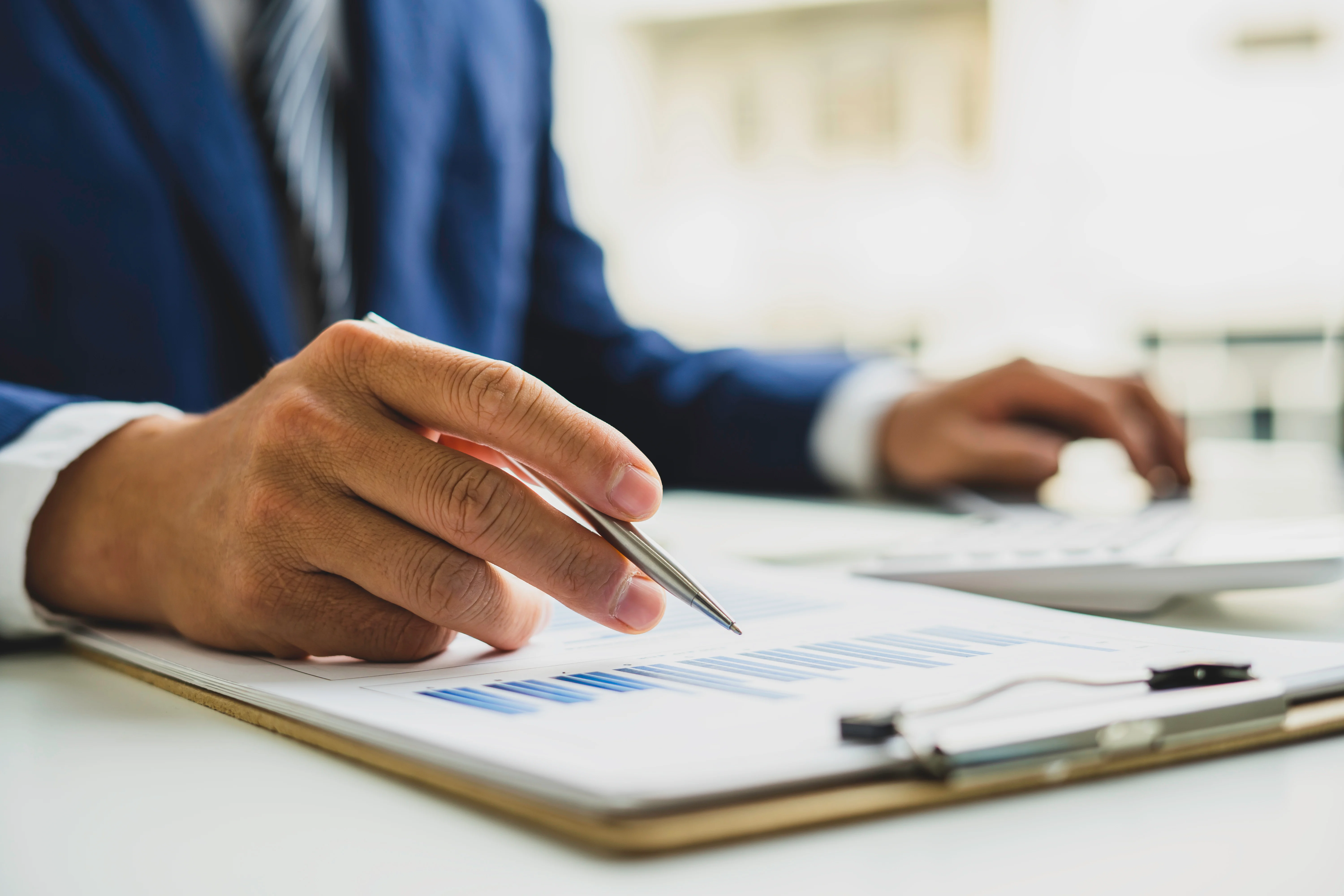 Een persoon in een pak wijst met een pen naar grafieken op een document terwijl hij met de andere hand een toetsenbord gebruikt.