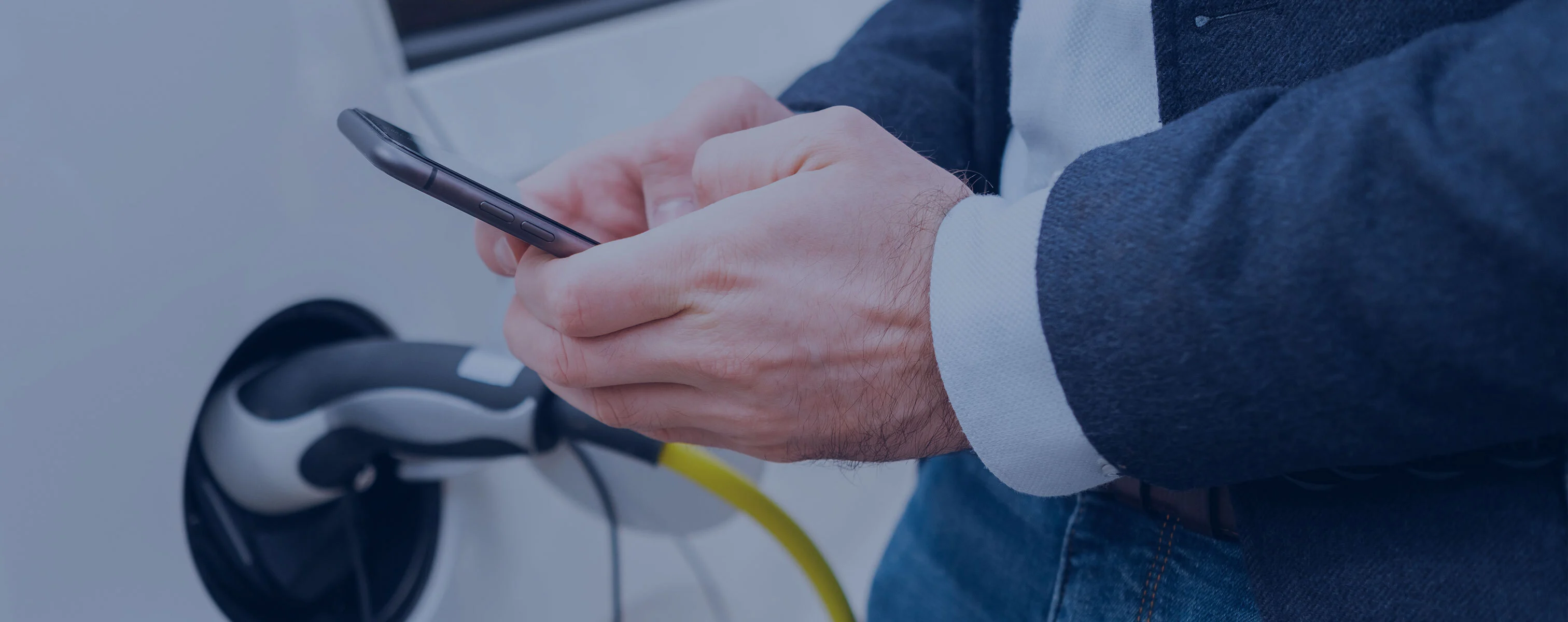Osoba trzyma smartfon w dłoni, podczas gdy samochód elektryczny jest ładowany za pomocą kabla ładującego.