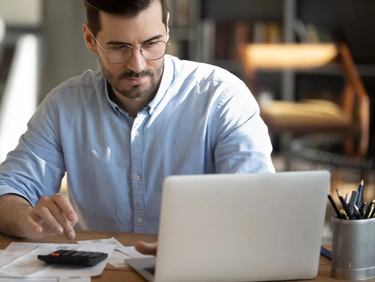 Mann arbeitet an einem Laptop und nutzt einen Taschenrechner, um Finanzdokumente zu überprüfen. Auf dem Tisch liegen verschiedene Papiere und eine Pflanze.