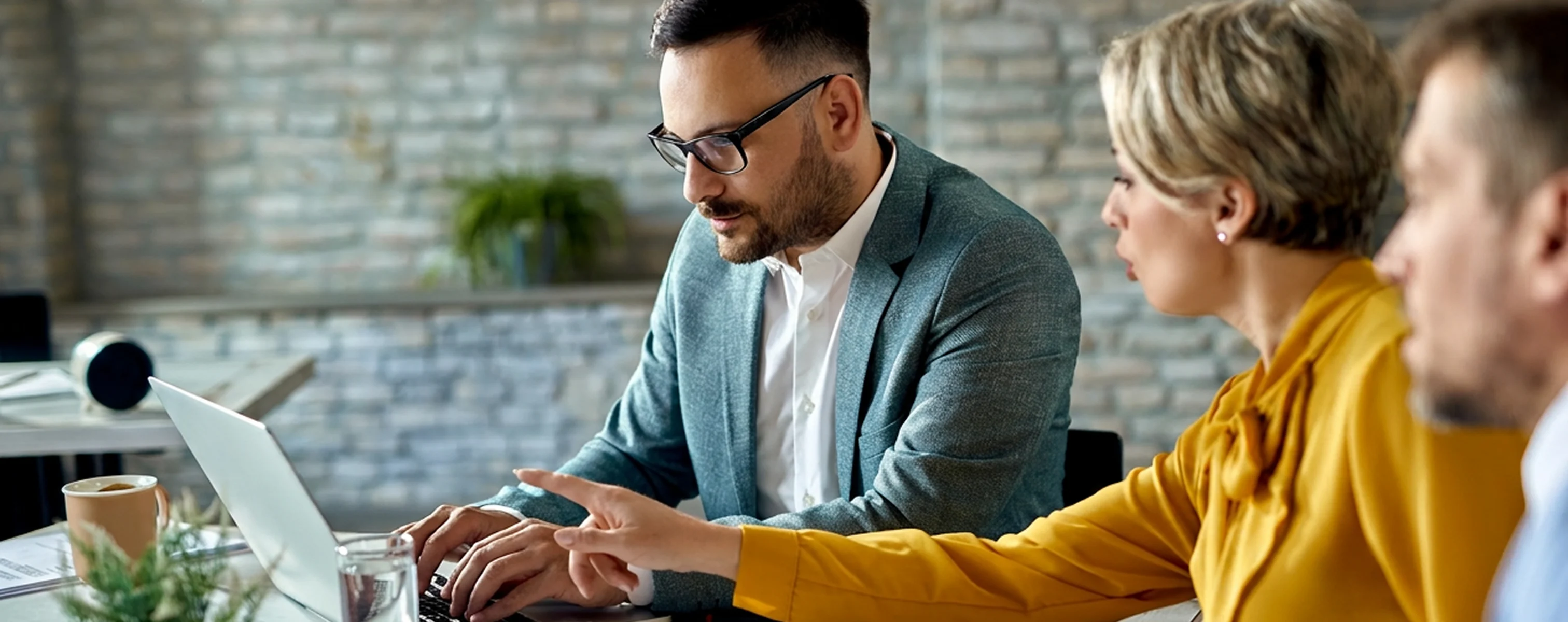 Drei Personen sitzen an einem Tisch und arbeiten gemeinsam an einem Laptop, während sie Dokumente durchgehen und etwas auf dem Bildschirm besprechen.