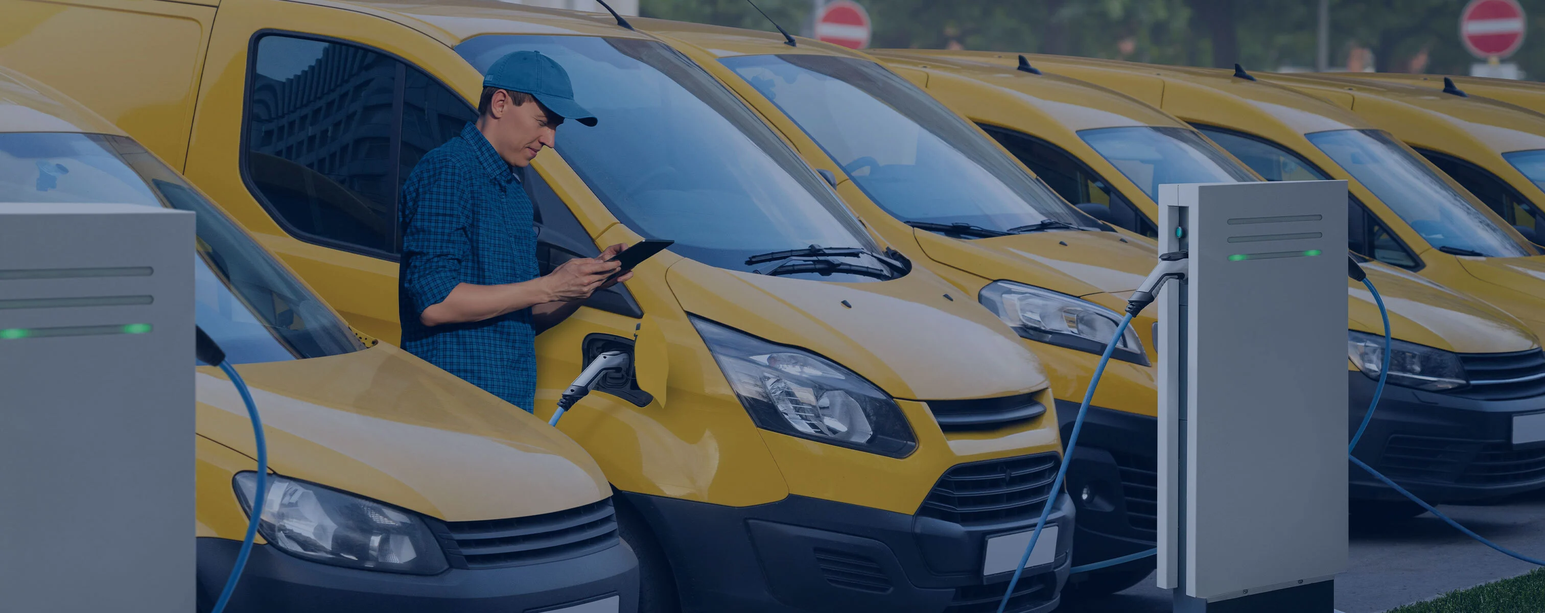 Une rangée de véhicules de livraison jaunes à une station de recharge électrique, tandis qu'un homme en chemise bleue et casquette de baseball vérifie le processus de recharge sur une tablette.