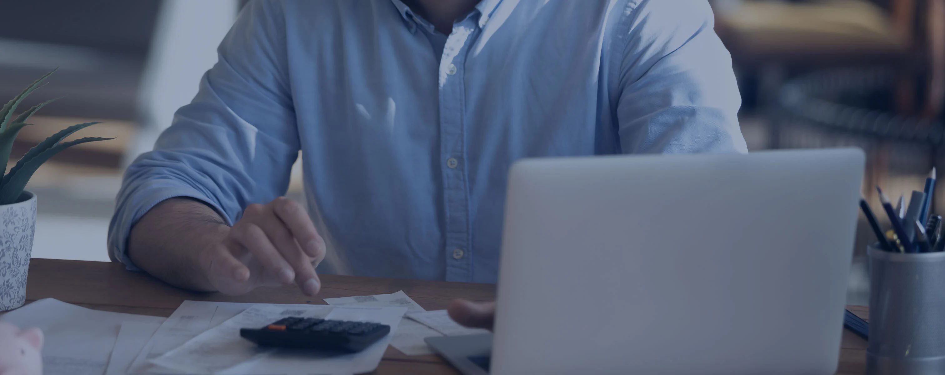 Mann arbeitet an einem Laptop und nutzt einen Taschenrechner, um Finanzdokumente zu überprüfen. Auf dem Tisch liegen verschiedene Papiere und eine Pflanze.