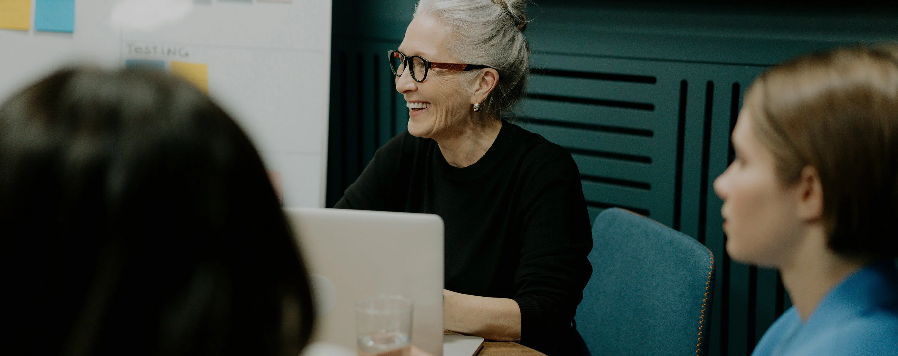 Eine ältere Frau lächelt während eines Meetings und schaut auf einen Laptop, während sie von Kollegen umgeben ist.