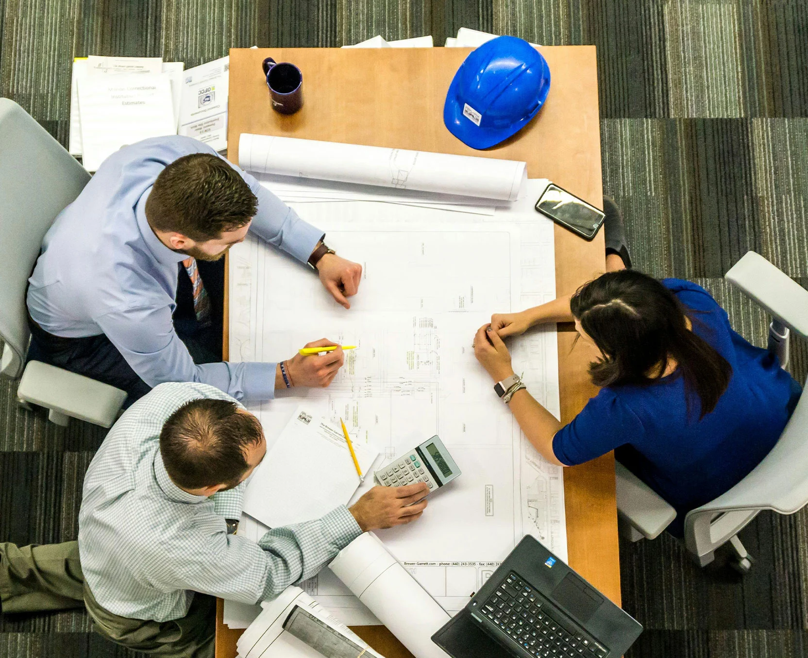 Trois employés travaillent autour d’une table avec des plans, un ordinateur portable et un calculatrice.