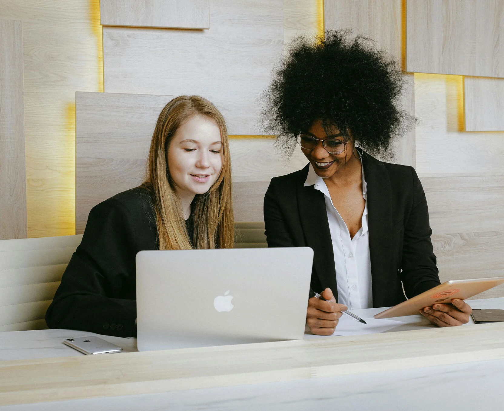 Zwei Frauen im Business-Outfit arbeiten gemeinsam lächelnd am Laptop.