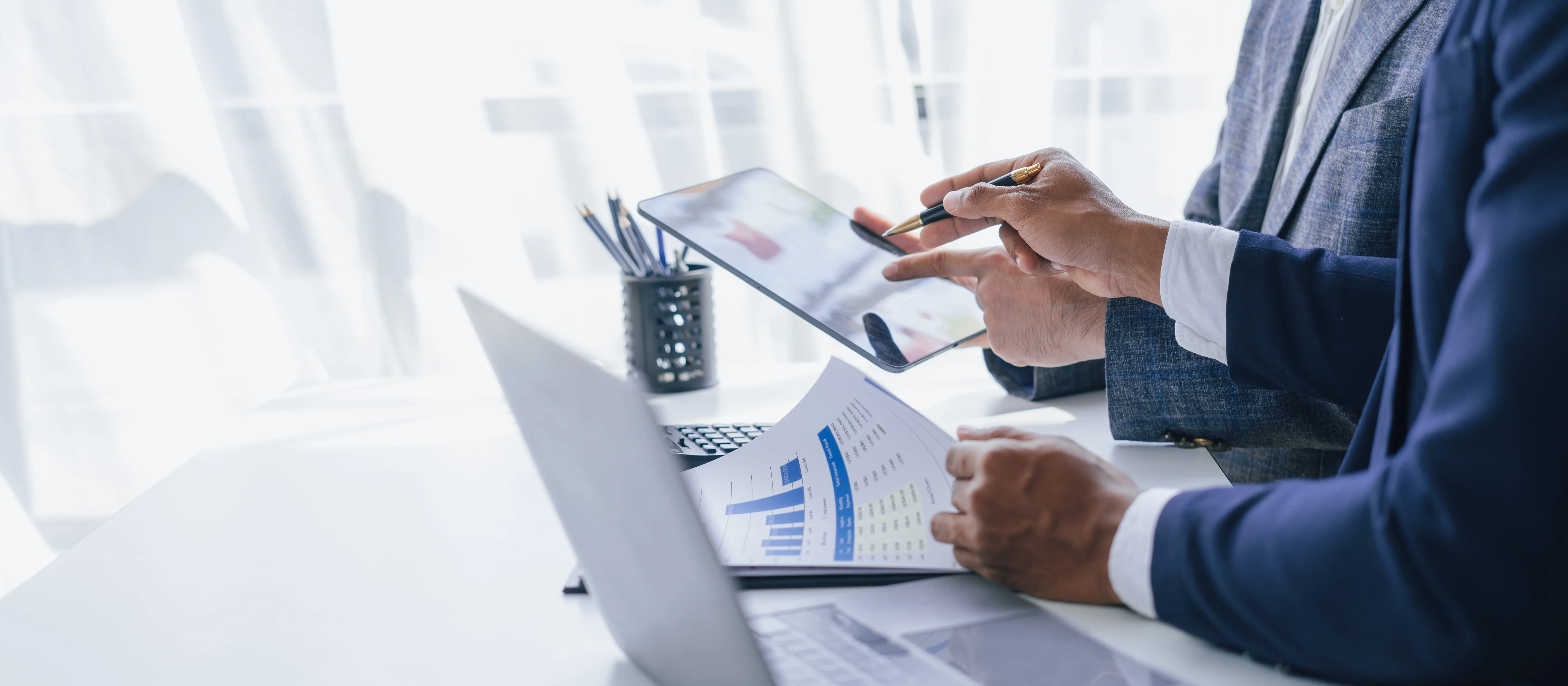 Zwei Geschäftspersonen in formeller Kleidung analysieren Daten. Eine Person zeigt mit einem Stift auf ein Tablet, während die andere ein Dokument mit Diagrammen und Statistiken hält