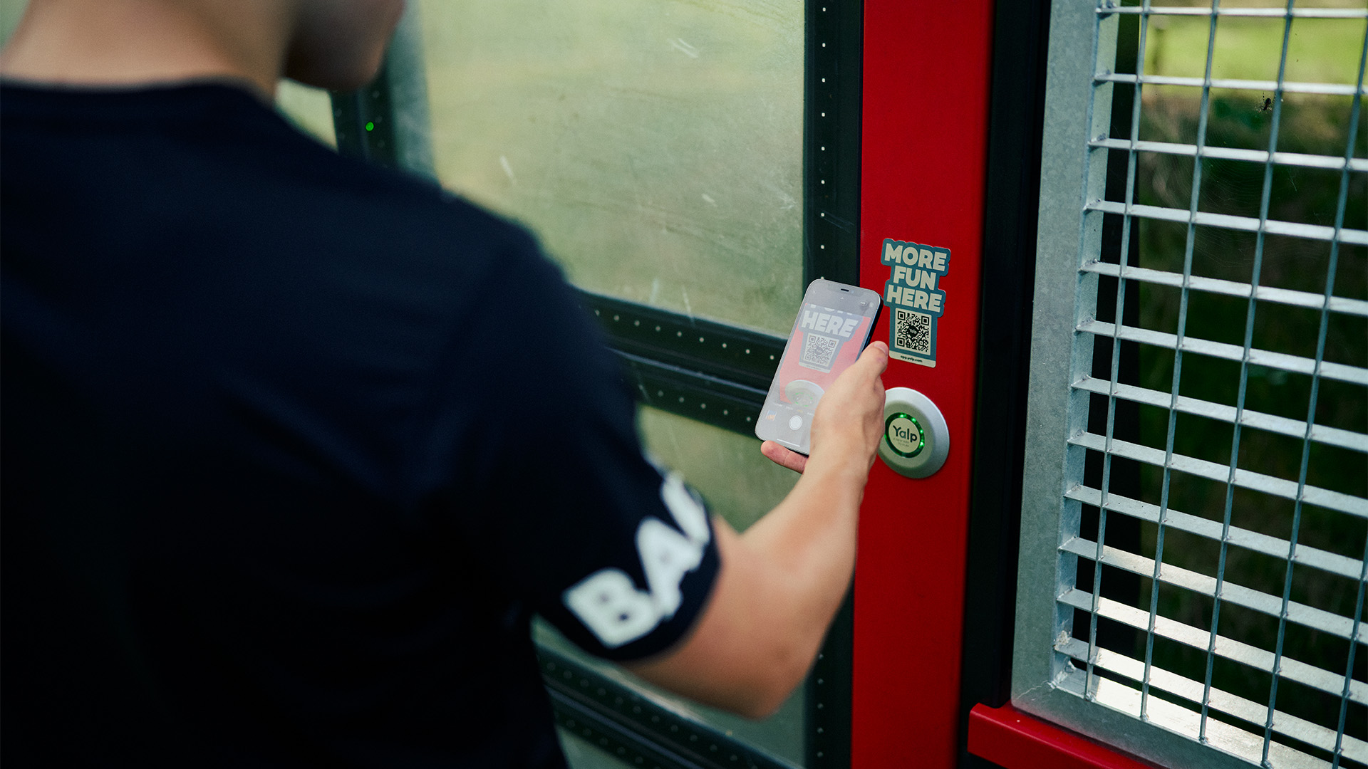 A person wearing a black shirt is scanning a QR code labeled "MORE FUN HERE" on a red door using a smartphone. The person is standing in front of the door, which has a metal grid and glass panel.