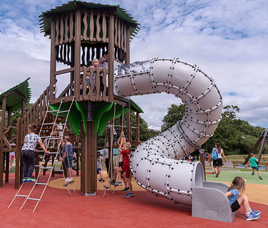 Children are playing on a playground with various structures, including a wooden tower with a spiral tube slide. Some children are climbing up ladders to reach the tower, while others are sliding down or playing on swings in the background under a partly cloudy sky.