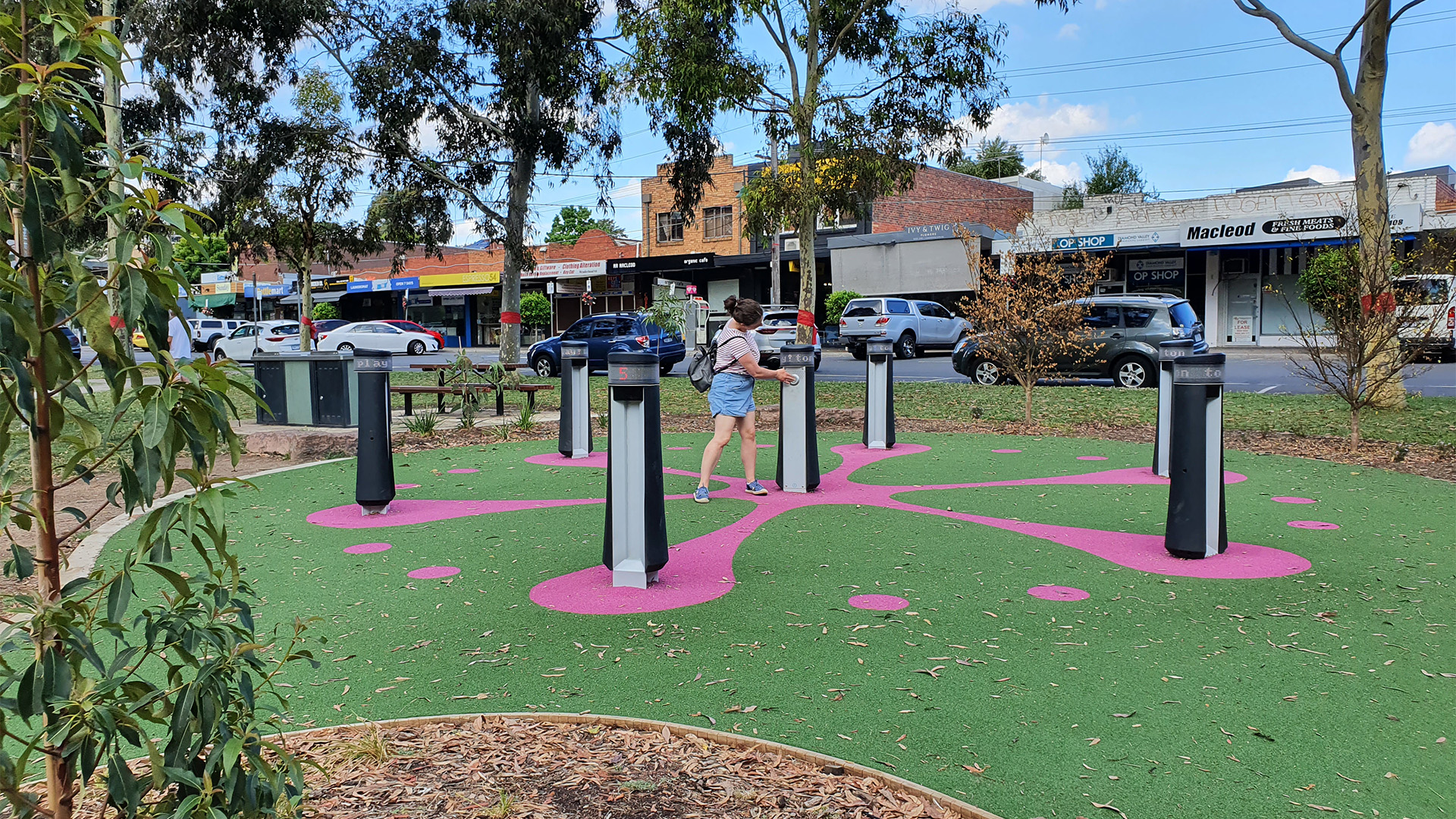 A person interacts with outdoor musical play equipment in a park with a colorful pink and green ground pattern. Shops and trees are visible in the background. The clear sky suggests it is daytime.