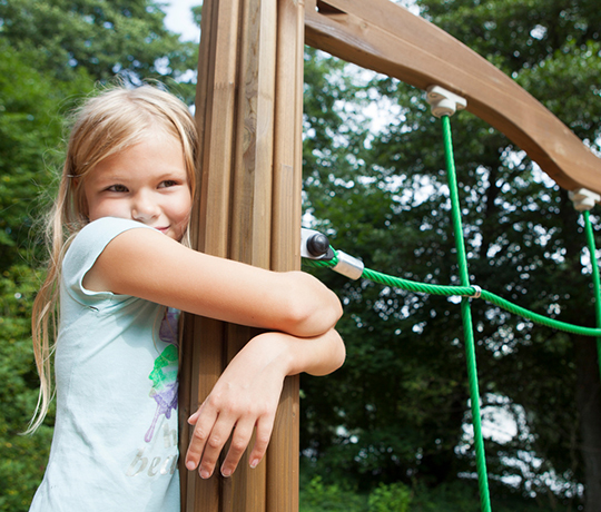 A young girl with blonde hair smiles and hugs a wooden post on a play structure. She is outdoors, surrounded by green trees, and is wearing a light-colored t-shirt. The play structure includes green ropes. The sun is shining, creating a bright and cheerful atmosphere.