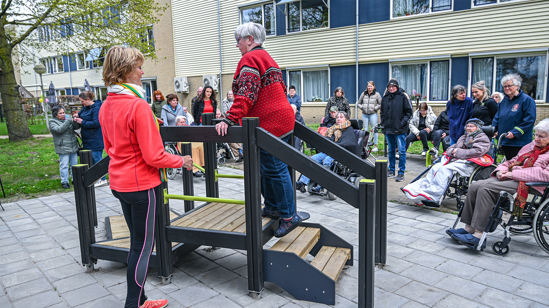 A group of people are gathered outdoors near a building, observing an older woman in a red sweater as she utilizes a small wooden bridge with assistance from another woman in a red jacket. Some individuals are seated in wheelchairs, and others are standing around.