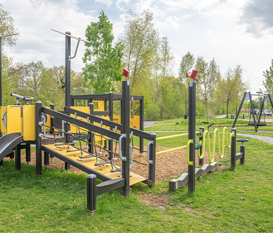 A playground featuring climbing equipment, ramps, and various obstacles made of yellow and black materials, set on grass and wood chip surfaces. Trees and greenery surround the area under a partly cloudy sky.