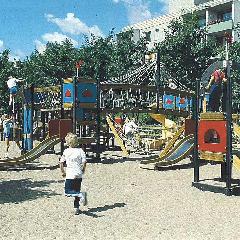 A playground featuring a large wooden play structure with slides, rope bridges, and climbing areas. Several children are playing, climbing, and running on and around the structure on a sandy ground. Trees and residential buildings are visible in the background.