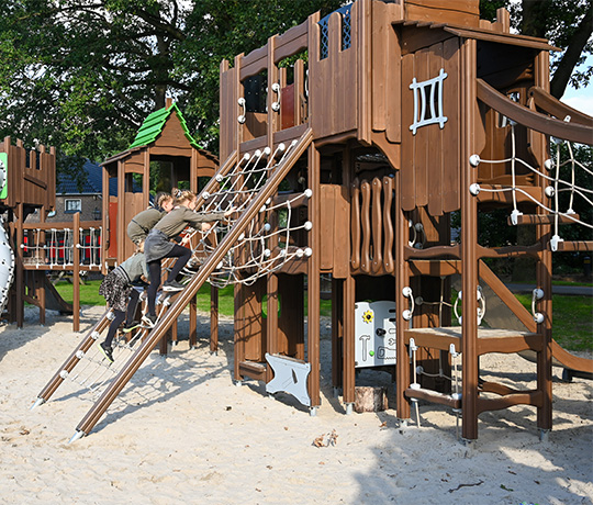 Two children are climbing a brown wooden play structure in a playground. The structure has various climbing elements, including a rope ladder, netting, and slides. The ground is covered in sand, and green grass and trees are visible in the background.