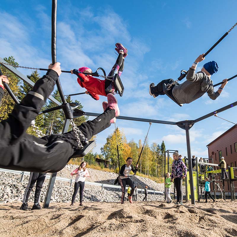 Children swing high in the air at a playground on a sunny day. The playground features multiple swings and play structures. Several kids are laughing and having fun, while adults and other children watch and enjoy the lively atmosphere around them.