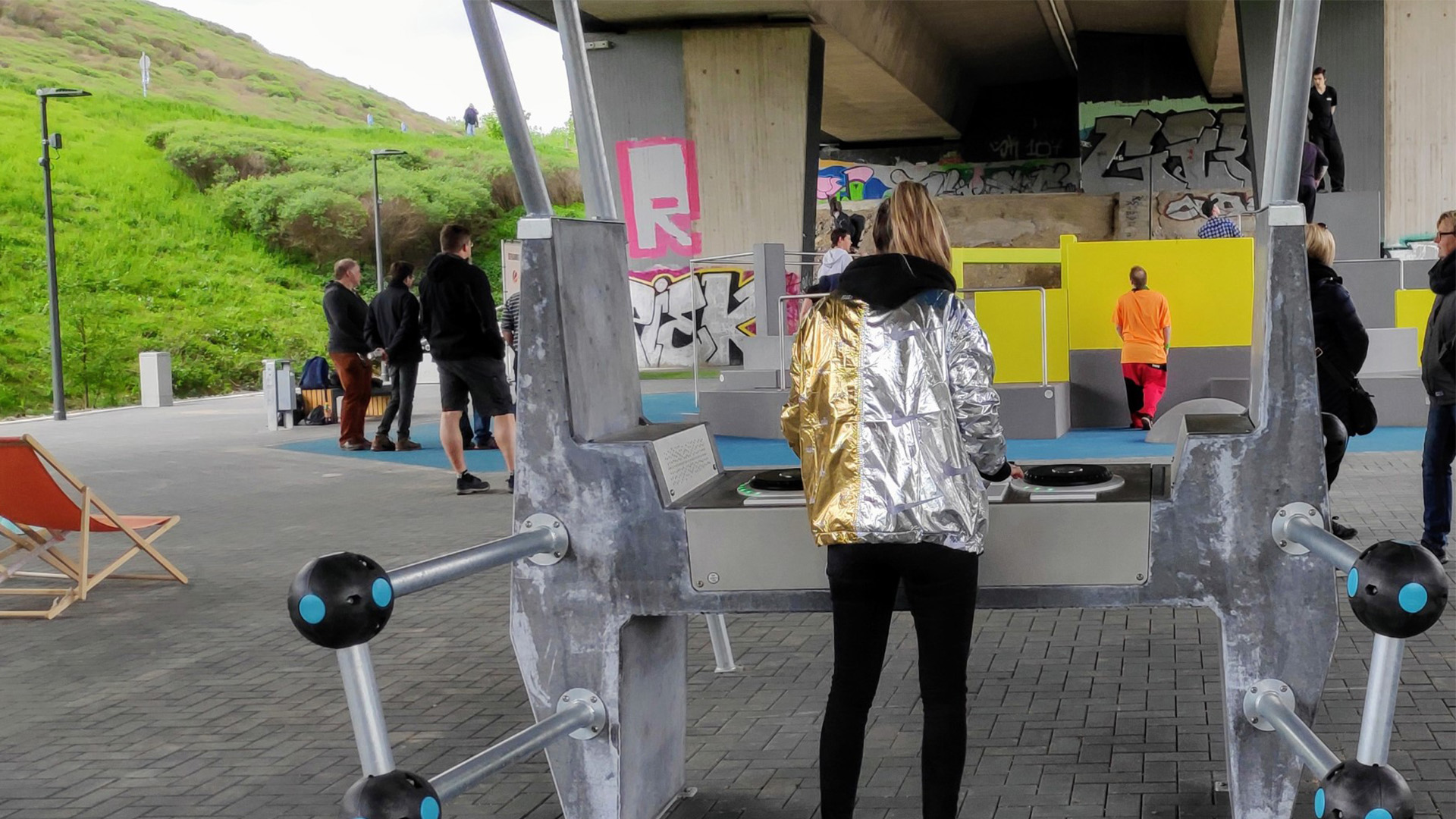 A person wearing a shiny, metallic jacket with gold and silver sleeves plays on an outdoor exercise machine beneath a bridge. Other individuals are seen in the background, and the area features colorful graffiti, lush green scenery, and modern seating.