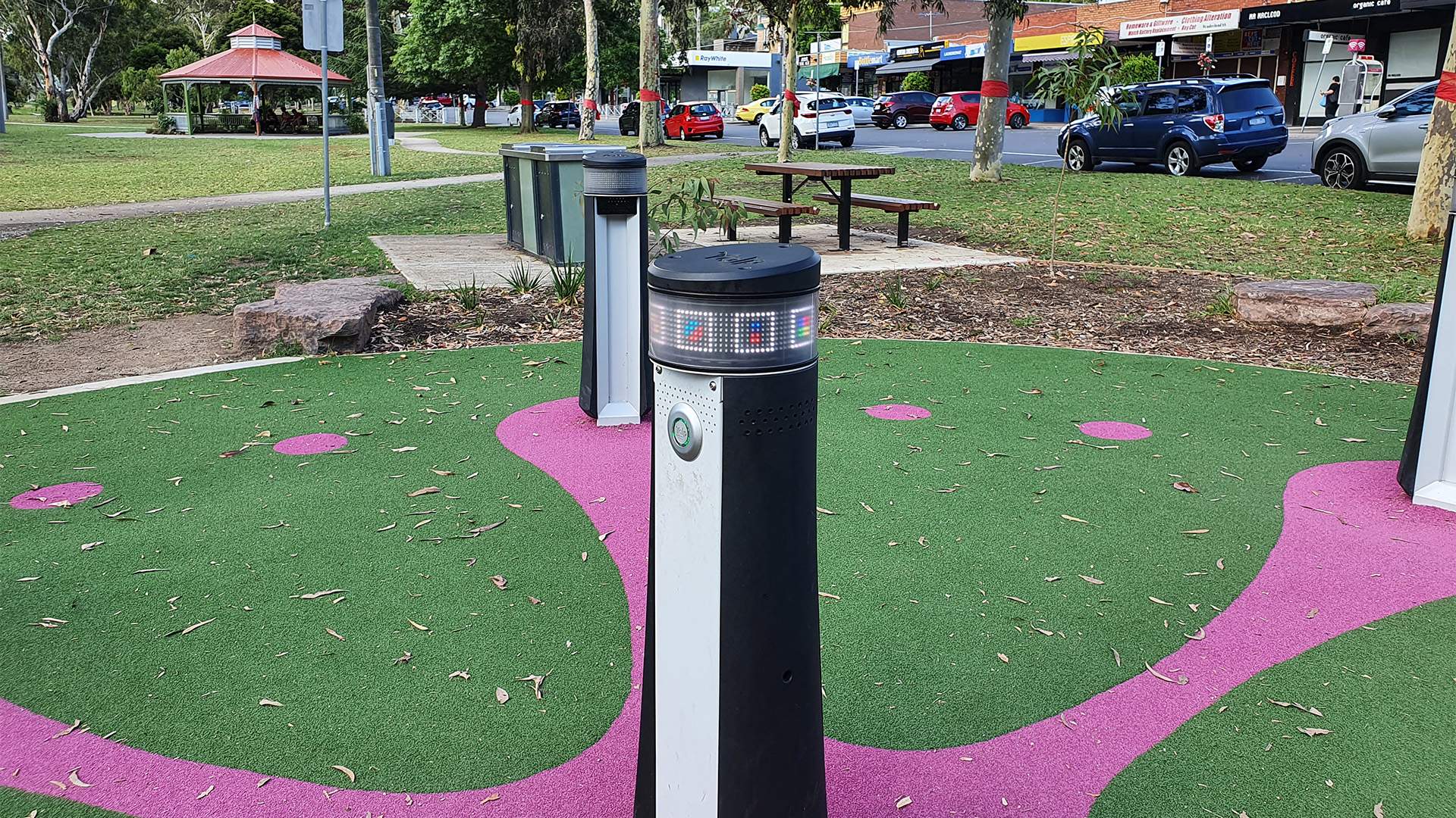 A park scene with colorful decorative bollards on a green and pink patterned surface. In the background, there are trees, a gazebo, benches, and a row of parked cars along a street with shops. The sky is clear and the setting appears calm.