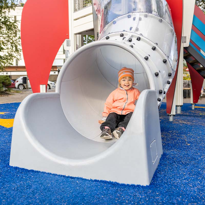 A young child wearing an orange jacket and hat exits a rocket-shaped playground slide and smiles. The surrounding playground features blue ground covering and colorful play structures in the background.