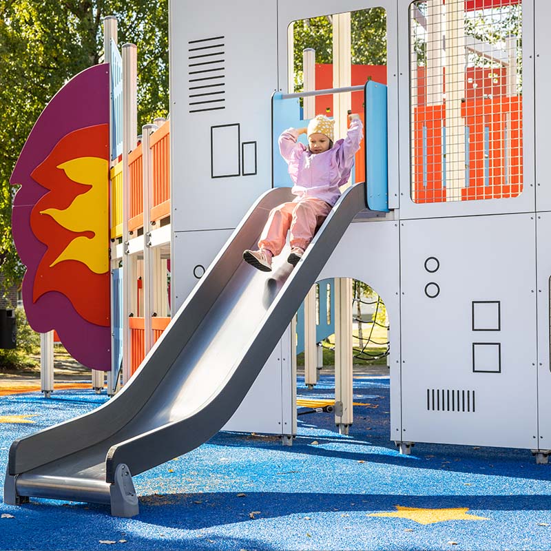A child in a pink outfit and yellow hat slides down a playground slide designed to look like part of a rocket ship. The playground features colorful components, including a rocket flame design, and is set on a blue surface with yellow star patterns. Trees can be seen in the background.
