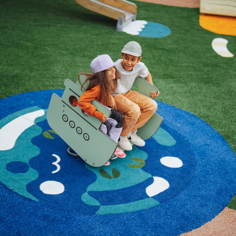Two children playing on a rocking Lappset Prime play equipment on a green and blue surface.