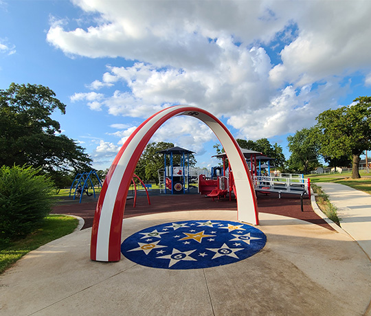 A patriotic-themed playground features a large arch with red, white, and blue stripes at the entrance. The ground beneath the arch displays yellow stars and a blue background. Various playground equipment is visible in the background against a partly cloudy sky.