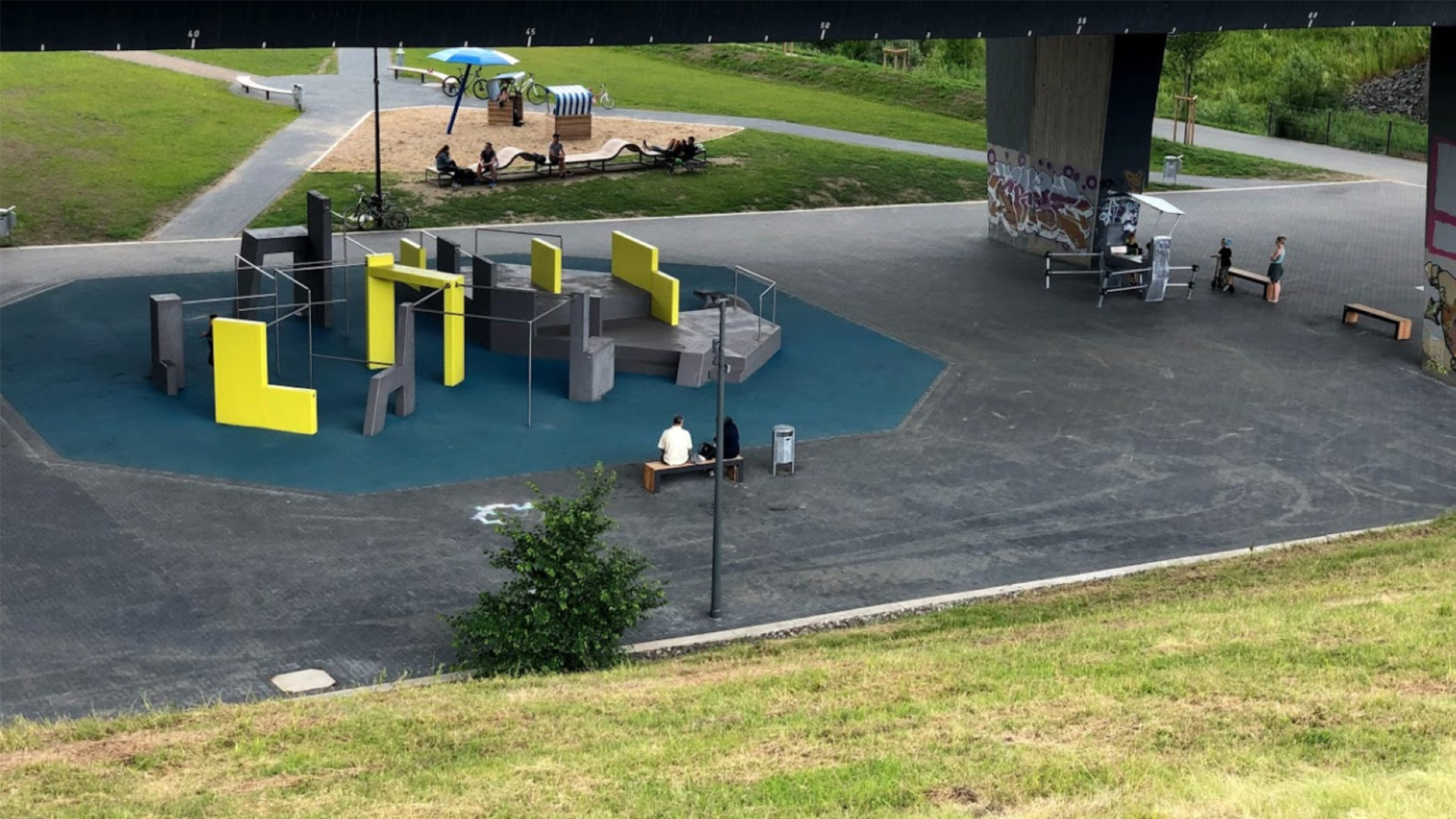 An outdoor parkour, play, and fitness area under a highway bridge featuring various climbing structures, benches, and graffiti. A park area with paths and playground equipment is visible in the background. A person sits on a bench, and another exercises.