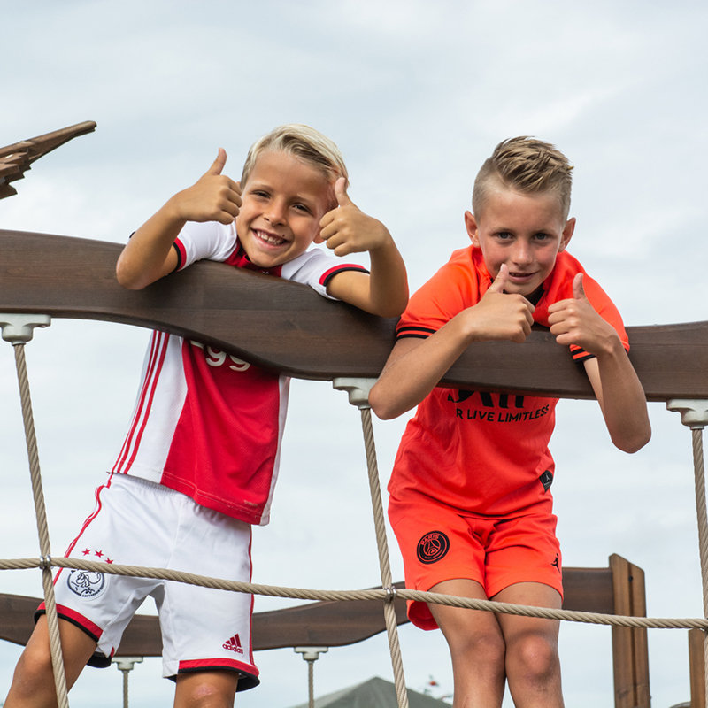 Two young boys stand on a wooden play structure, smiling and giving thumbs up. The boy on the left wears a white and red sports uniform, while the boy on the right wears an orange sports uniform. The sky is cloudy in the background.