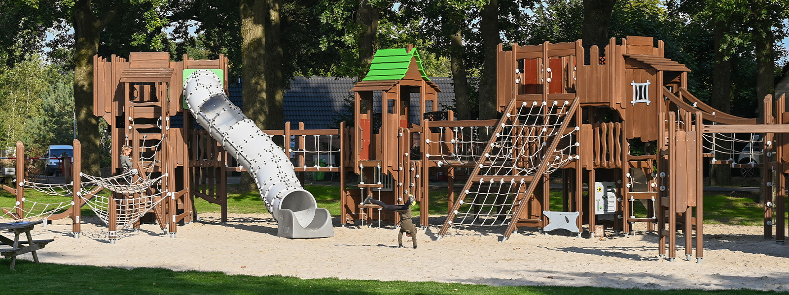 A large, outdoor playground featuring various wooden play structures. There are climbing nets, slides, and bridges, all set on a sandy surface. Trees provide shade in the background, and the playground is surrounded by lush green grass.